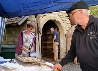 Das Bild zeigt einen Mann und eine Frau an einem Verkaufsstand. Zwischen ihnen liegt eine große rechteckige Pizza.