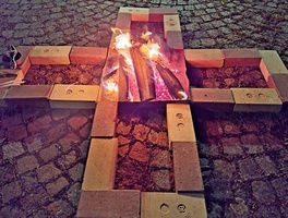 Das Bild zeigt ein Feuer, das in Form eines Kreuzes aus Ziegelsteinen auf einem Kopfsteinpflaster angeordnet ist. Die Flammen brennen in der Mitte des Kreuzes. Neben dem Kreuz liegt ein Werkzeug auf dem Boden.