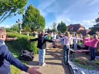 Auf dem Friedhof in Püssensheim stehen Menschen und machen eine weite Bewegung mit den Händen.
