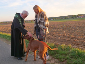 Auf dem Bild streichelt ein Pater einen braunen mittelgroßen Hund am Kopf. Hinter dem Hund steht offenbar seine Besitzerin.