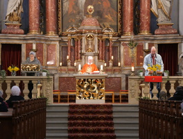 Das Bild zeigt den Altarbereich einer Kirche. In der Mitte steht ein Priester in orangefarbener Robe, links und rechts von ihm stehen zwei Helfer. Im Hintergrund befinden sich aufwendig gestaltete Säulen und Statuen.