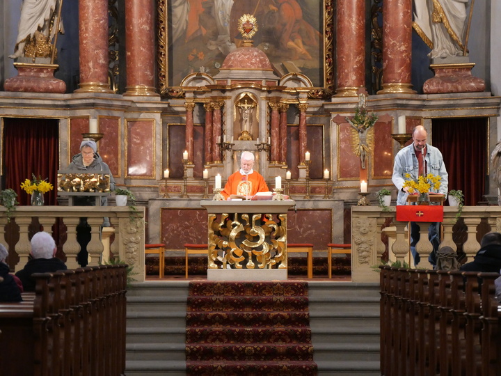 Das Bild zeigt den Altarbereich einer Kirche. In der Mitte steht ein Priester in orangefarbener Robe, links und rechts von ihm stehen zwei Helfer. Im Hintergrund befinden sich aufwendig gestaltete Säulen und Statuen.
