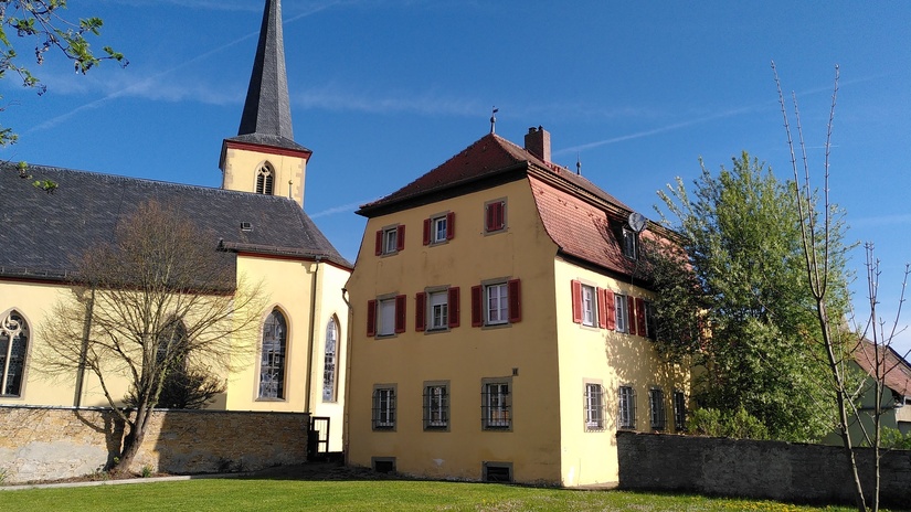 Das Bild zeigt eine gelbe Kirche mit hohem, spitzem Turm und ein angrenzendes Gebäude mit roten Fensterläden unter klarem blauem Himmel. Grünes Rasenstück vorne.