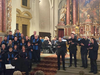 Das Bild zeigt einen Chor in einer Kirche. Die Mitglieder sind schwarz gekleidet und halten Notenblätter. Zwei Dirigenten stehen vorne und leiten den Chor. Im Hintergrund sieht man einen Altar mit einem Gemälde und einem Kreuz.