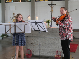 Zwei Musikerinnen spielen in einer Kirche mit zwei Geigen. Notenständer, Kerzen und ein Kruzifix sind im Hintergrund sichtbar.