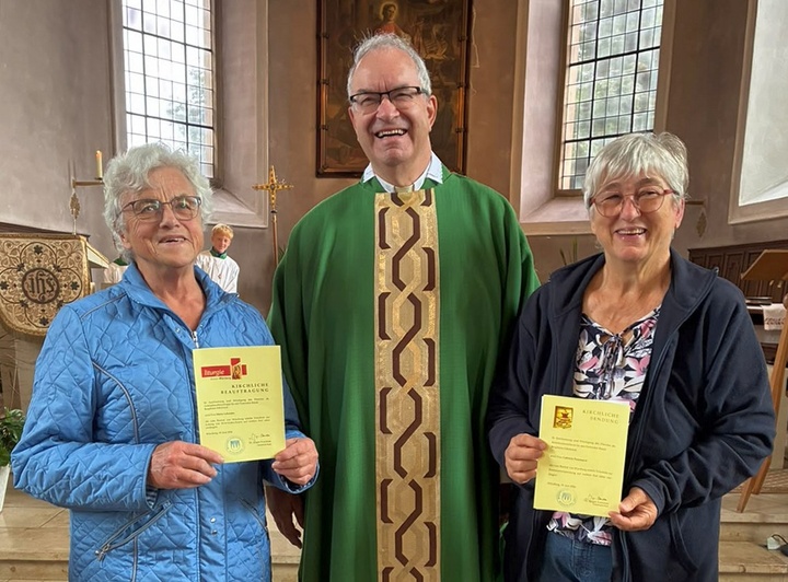 Das Bild zeigt zwei Frauen mit Urkunden in der Hand. In der Mitte ist Pfarrer Helmut Rügamer. Das Bild wurde in einer Kirche aufgenommen.