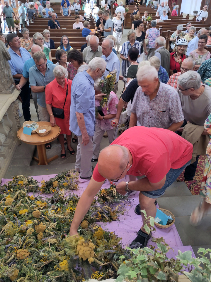 Das Bild zeigt das Innere einer Kirche. Man sieht viele Menschen in den Bänken und im Gang. Im Vordergrund liegen Blumen auf dem Altarstufen. Manche werden gerade aufgehoben.