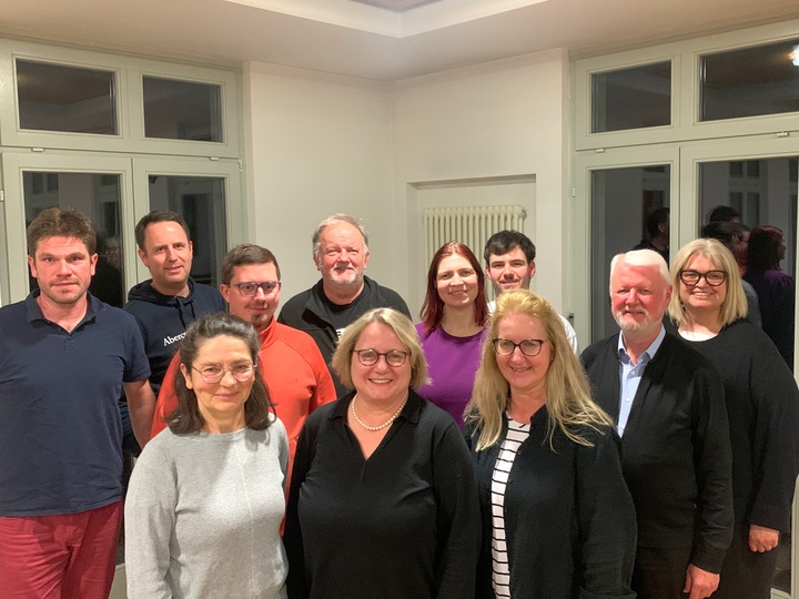 Das Bild zeigt eine Gruppe von elf Personen, die in einem hellen Raum mit großen Fenstern und weißen Wänden posieren. Sie stehen in zwei Reihen. Die Personen sind lässig bis leger gekleidet und lächeln in die Kamera.