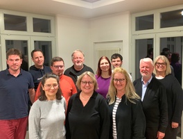 Das Bild zeigt eine Gruppe von elf Personen, die in einem hellen Raum mit großen Fenstern und weißen Wänden posieren. Sie stehen in zwei Reihen. Die Personen sind lässig bis leger gekleidet und lächeln in die Kamera.