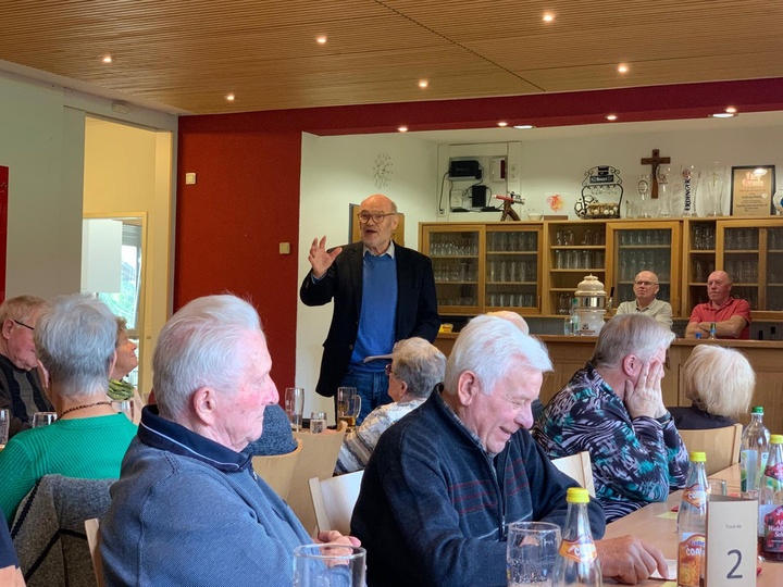 Blick auf mehrere ältere Menschen, die an verschiedenen Tischen sitzen; vorne steht ein Mann und erzählt etwas.