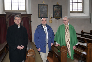 Drei Männer stehen in Kirchenbänken. Hinter ihnen an der Wand hängen zwei der Kreuzwegbilder.