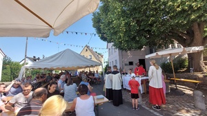 Das Bild zeigt in der Mitte den Altar, um den sich der Priester und die Ministrant:innen versammeln. In der linken Hälfte des Bildes sitzen Menschen an Biertischen.