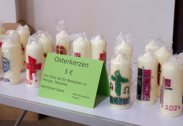 Verschiedene Osterkerzen stehen auf einem Tisch. Davor ist ein grünes Schild. Darauf steht, wofür der Erlös der Aktion gedacht ist. 