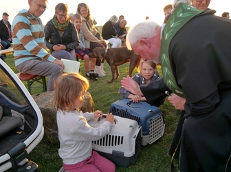 Das Bild zeigt einen Pater, der sich zu Kindern hinunterbeugt und eine Segensgeste macht. Zwischen den Kindern sind zwei Tansportboxen für kleine Tiere zu sehen.