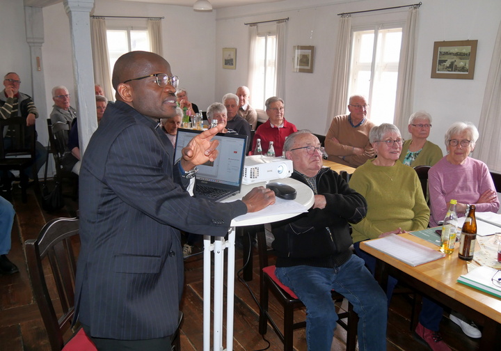 Ein Mann in einem dunklen Anzug hält eine Präsentation vor einer Gruppe von älteren Menschen. Er steht neben einem Tisch mit einem Laptop und einem Projektor. Die Zuhörer sitzen an Tischen und hören aufmerksam zu.