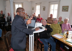 Ein Mann in einem dunklen Anzug hält eine Präsentation vor einer Gruppe von älteren Menschen. Er steht neben einem Tisch mit einem Laptop und einem Projektor. Die Zuhörer sitzen an Tischen und hören aufmerksam zu.