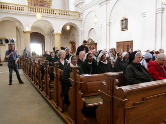 Das Bild zeigt eine Gruppe von Menschen, die in einer Kirche auf Holzbänken sitzen. Die meisten tragen Nonnentracht oder religiöse Kleidung. Die Kirche hat hohe weiße Wände, Bögen und dekorative Elemente. Ein Lautsprecherträger steht links im Vordergrund.