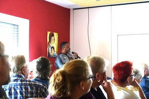 Das Foto ist vom Sitzplatz in der Versammlung aufgenommen. Man sieht den Referenten im Hintergrund. Alle blicken zur Präsentation an der Wand.