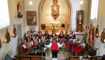 Eine Musikkapelle mit Blasinstrumenten sitzt im vorderen Bereich einer Kirche. Ein Mann dirigiert.