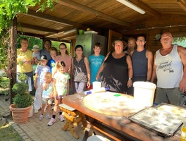 Etwa 12 Erwachsene und Kinder stehen nebeneinander und lachen in die Kamera, vor ihnen liegt das Zubehör zum Pizza backen.