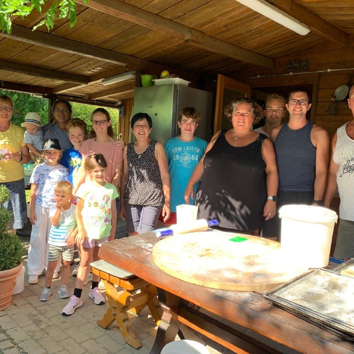 Etwa 12 Erwachsene und Kinder stehen nebeneinander und lachen in die Kamera, vor ihnen liegt das Zubehör zum Pizza backen.