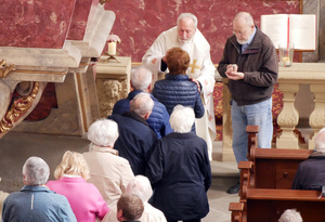 Beim Blick von der Empore auf einen segnenden Priester. In einer Reihe stehen ältere Menschen und warten auf ihre Krankensalbung.