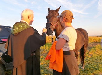 Auf diesem Foto steht ein braunes Pferd in der Mitte. Links streichelt ein Pater seinen Kopf. Rechts ist offenbar die Pferdebesitzerin. Es ist ein sonniges Bild im Abendrot.