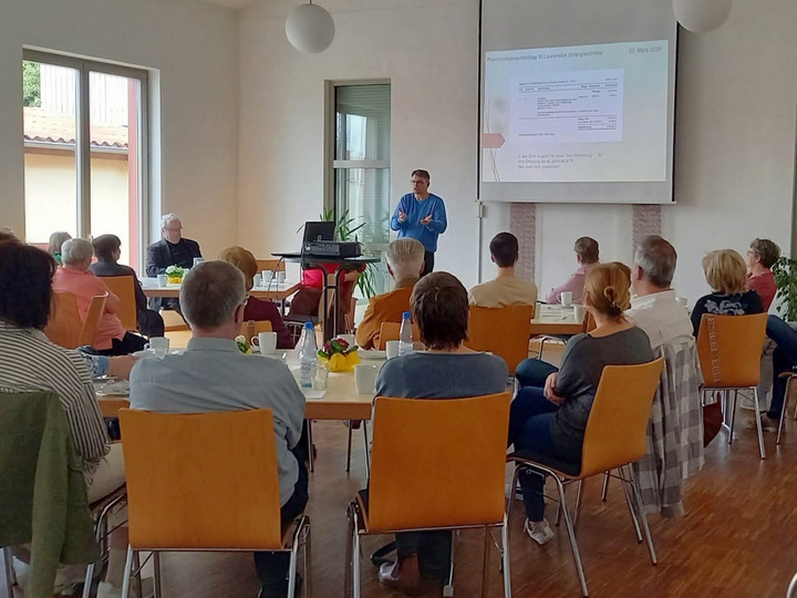 Ein schönes helles Bild mit Blick in das Pfarrheim. Auf Stühlen sitzen die Menschen und schauen nach vorn zum Redner.