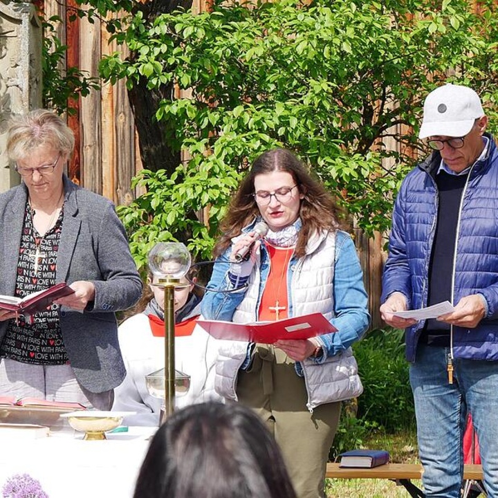 Drei Personen stehen mit Mappen und Mikrofon vor anderen und tragen Gebete vor.