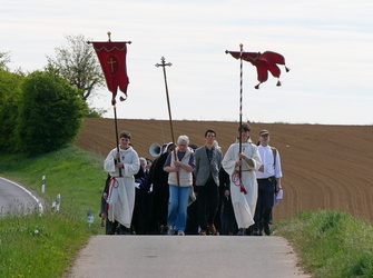 Das Bild zeigt eine Gruppe von Menschen, die auf einer Landstraße gehen. Einige tragen religiöse Gewänder und halten Banner mit Kreuzen. Die Landschaft ist ländlich mit Feldern und Grünflächen.