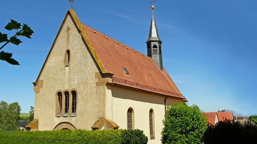 Die Abbildung zeigt eine kleine, historische Kirche mit rotem Ziegeldach, einem spitzen Turm und Kreuz. Sie ist von grünen Hecken umgeben und steht unter blauem Himmel.