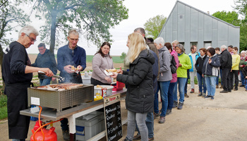 An einem Grill auf der linken Bildseite werden Bratwürste gegrillt. Rechts stehen Menschen in einer Schlange an.