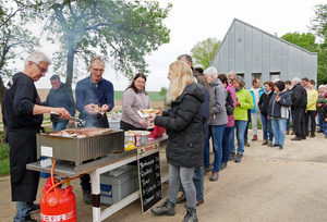 An einem Grill auf der linken Bildseite werden Bratwürste gegrillt. Rechts stehen Menschen in einer Schlange an.