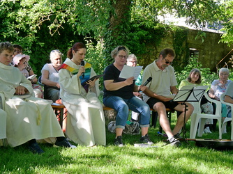 Auf dem Bild sieht man eine Gruppe Menschen im Freien sitzen, die an einer Veranstaltung teilnehmen. Es ist ein sonniger Tag mit viel Grün.