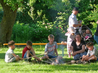 Eine Gruppe Menschen sitzt im Grünen um ein kleines Feuer. Erwachsene und Kinder bereiten gemeinsam etwas am Spieß über der Flamme zu. Bäume spenden Schatten.