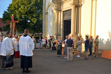 Auf dem Parkplatz vor der Kirche stehen rechts die Musikanten und spielen ein Lied. Der Platz liegt in der Abendsonne.