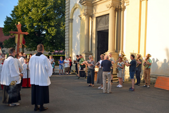 Auf dem Parkplatz vor der Kirche stehen rechts die Musikanten und spielen ein Lied. Der Platz liegt in der Abendsonne.