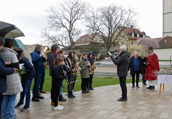 Die Musikkapelle spielt ein Lied. Rechts stehen der Pfarrer und der Küster. Es ist ein regnerischer Tag. Schirme sind aufgespannt.