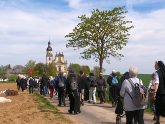 Das Bild zeigt eine Gruppe von Menschen, die auf einem Feldweg in Richtung einer Kirche mit einem hohen Turm gehen. Einige tragen Rucksäcke und religiöse Kleidung. Ein Baum steht rechts im Vordergrund, und der Himmel ist klar mit einigen Wolken.