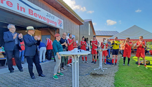 Pfarrer Rügamer, Bischof Franz Jung und eine Gruppe in roten Fußballtrikots stehen um kleine Tische und klatschen. Auf einem Tisch steht der Pokal.