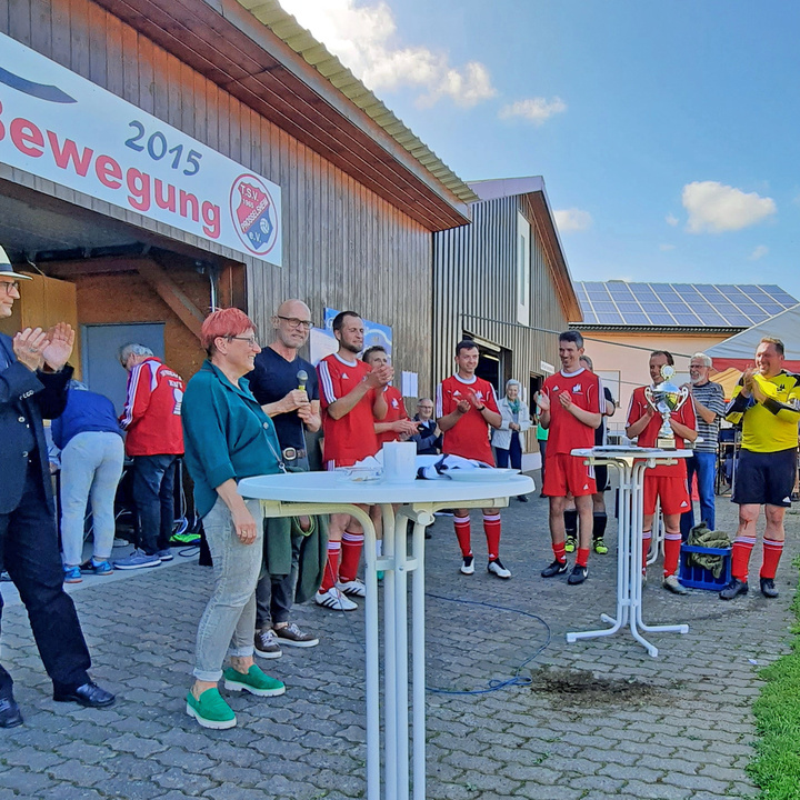 Pfarrer Rügamer, Bischof Franz Jung und eine Gruppe in roten Fußballtrikots stehen um kleine Tische und klatschen. Auf einem Tisch steht der Pokal.