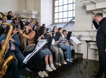Das Bild zeigt ein Orchester in einem hellen Raum mit großen Fenstern. Es sind verschiedene Blasinstrumente wie Flöten, Saxophone und Tubas zu sehen. Ein Dirigent steht rechts und leitet die Musikerinnen und Musiker an.