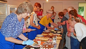 Auf einem langen Tisch stehen viele Kuchen und Torten. Frauen verteilen Stücke davon auf Teller.