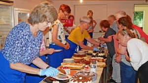 Auf einem langen Tisch stehen viele Kuchen und Torten. Frauen verteilen Stücke davon auf Teller.