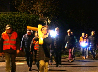 Auf dem Bild sind mehrere Personen nachts auf einer Straße zu sehen. Eine Person trägt ein großes, gelbes Kreuz, während andere mit Taschenlampen leuchten. Einige tragen Warnwesten. Es ist dunkel, mit Beleuchtung durch Taschenlampen und Straßenlaternen.