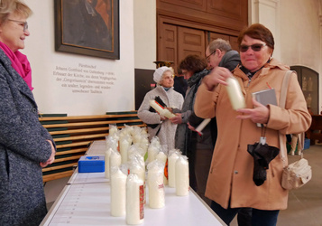 Auf einem Tisch in der Mitte stehen Osterkerzen. Frau Altenhöfer steht links hinter dem Tisch. Rechts vom Tisch schaut sich eine Frau eien Kerze an.