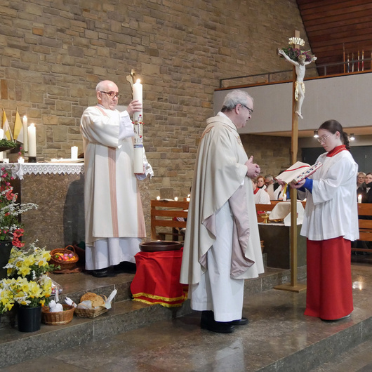Der Diakon hält die brennende Osterkerze hoch. Vor ihm stehen der Pfarrer und eine Ministrantin.