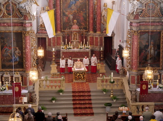 Das Bild zeigt den Innenraum einer prächtig geschmückten Kirche während einer Messe. Im Mittelpunkt steht ein Geistlicher in einem weißen Gewand und roter Stola. Die Kirche ist mit großen Gemälden, goldenen Verzierungen und Kerzenleuchtern ausgestattet. Ein roter Teppich führt zu einem Altar, der mit Kerzen und Blumen geschmückt ist.