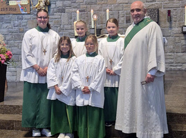In der Kirche stehen fünf Mädchen in weißen Chorhemden mit grünen Schärpen und ein lächelnder Geistlicher in weiß-grüner Robe vor einer Steinwand. Feierliche Atmosphäre.