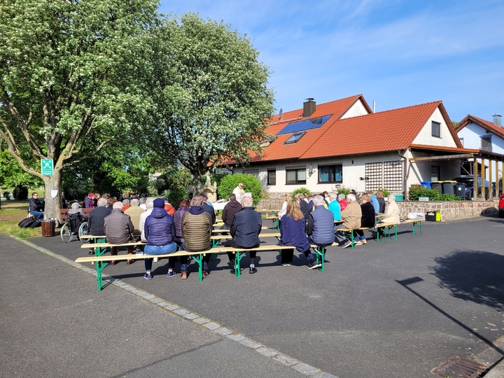 Mehrere Menschen sitzen auf Bierbänken unter einem Baum und feiern Gottesdienst.